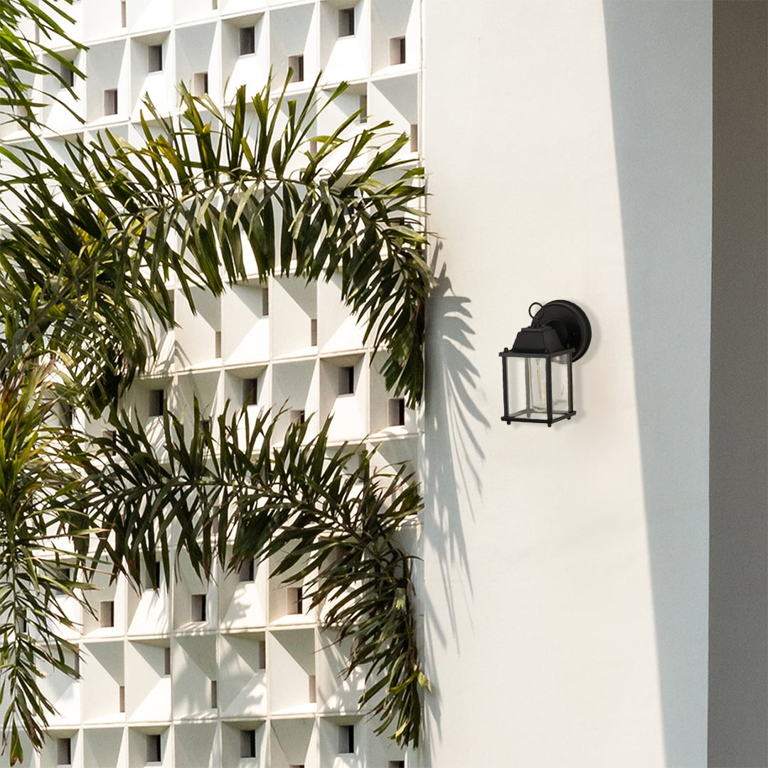 A modern outdoor wall light placed on a bright exterior wall surrounded by tall green foliage showing the fixture in a natural daylight setting with no light on.