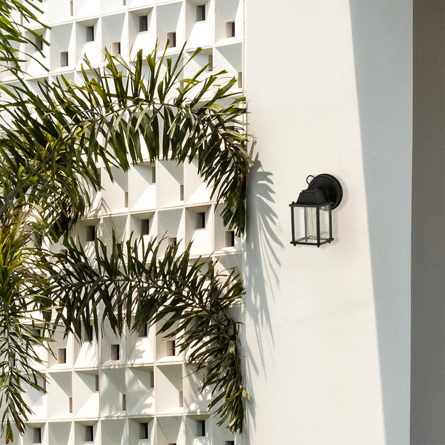 A modern outdoor wall light placed on a bright exterior wall surrounded by tall green foliage showing the fixture in a natural daylight setting with no light on.