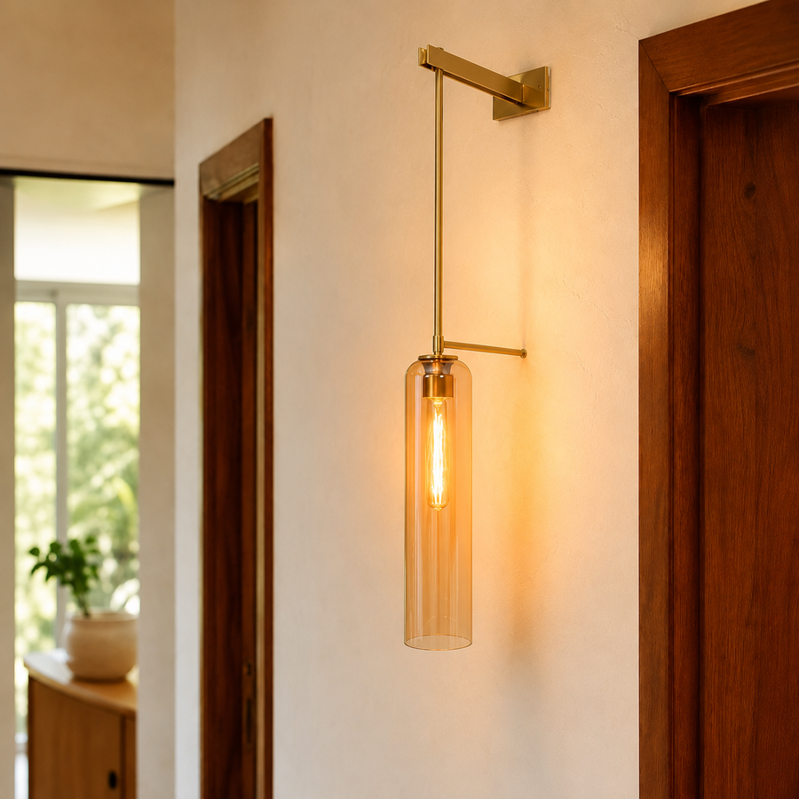 A modern wall sconce with a vertical clear glass cylinder shade and a warm glowing Edison bulb, mounted on a beige wall in a hallway with wooden doorframes.