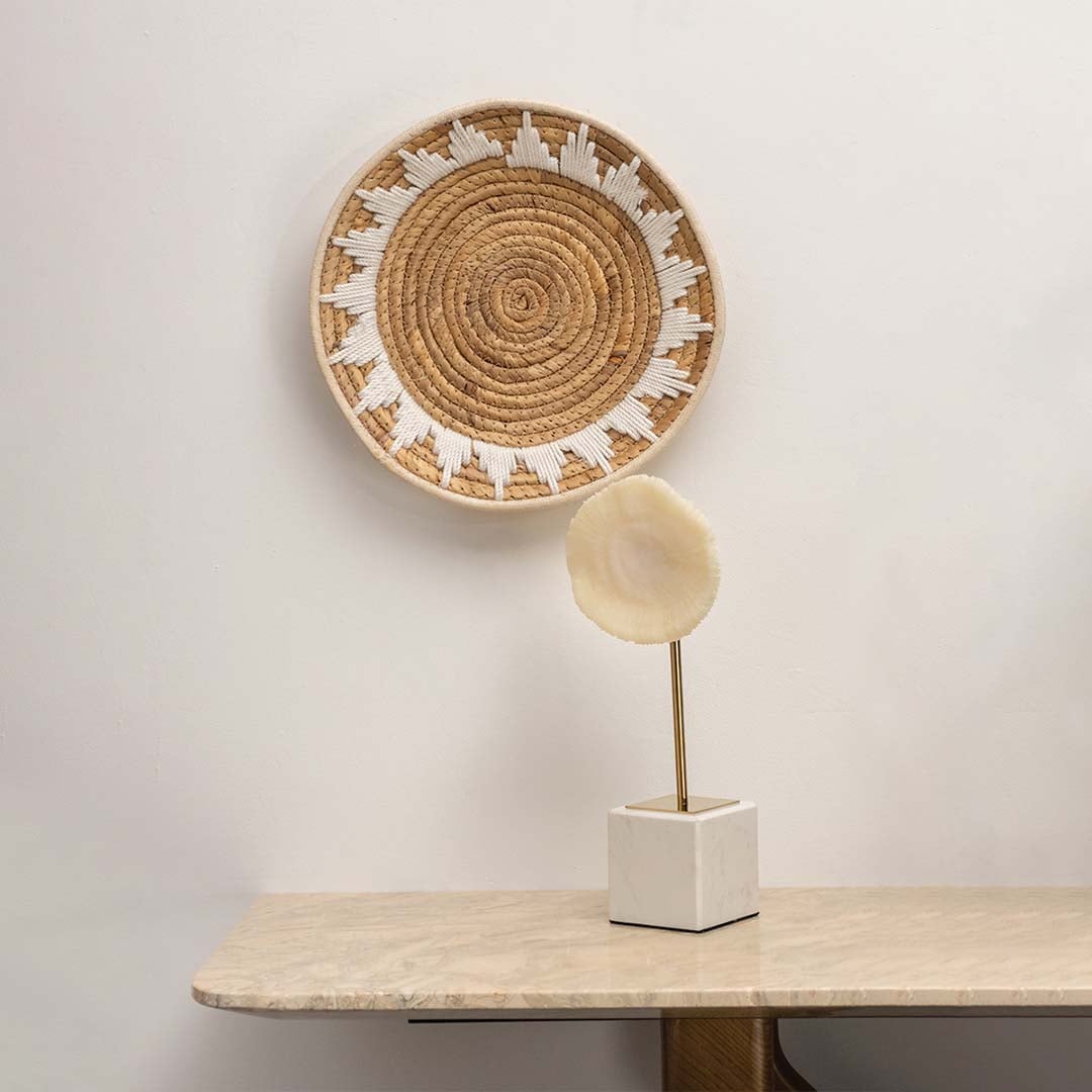 A natural woven wall plate with white embroidered pattern hanging on a light wall above a marble table styled with a modern decorative piece for a warm lifestyle look.