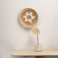Medium beige and white woven wall basket hanging on a white wall above a marble console table with a sculpture for boho home decor styling.