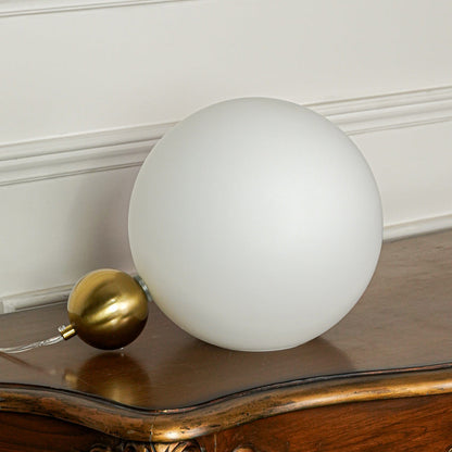 Contemporary matte white globe table lamp with brass finish base positioned on wooden table with neutral wall background ideal for modern lighting setup.