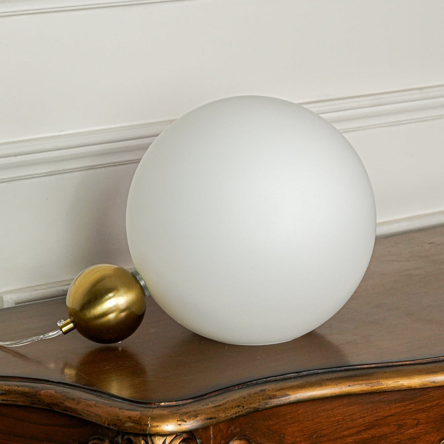 Contemporary matte white globe table lamp with brass finish base positioned on wooden table with neutral wall background ideal for modern lighting setup.