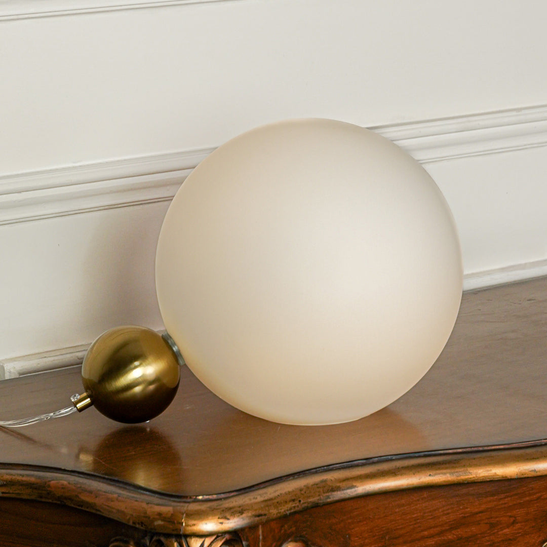 Modern white frosted glass globe table lamp with gold metal base placed on wooden surface against textured white wall background for elegant decor.