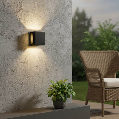 Modern black square outdoor up down wall light casting architectural glow on grey stucco wall near wicker chair and plant.