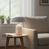 Table lamp styled on wooden side table beside soft armchair creating cozy ambient glow with white dome shade and smooth base for relaxed reading corners.