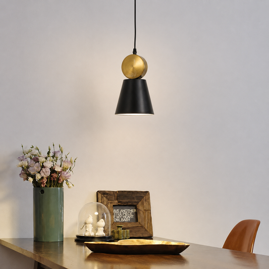 A modern pendant light with a black cone shade and a gold spherical accent hanging above a wooden table in a styled interior setting.