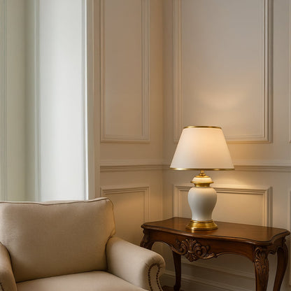 White ceramic table lamp with golden base and tapered fabric shade adding warm lighting to traditional decor.
