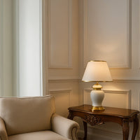 White ceramic table lamp with golden base and tapered fabric shade adding warm lighting to traditional decor.