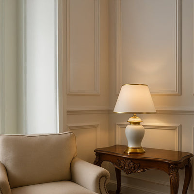 White ceramic table lamp with golden base and tapered fabric shade adding warm lighting to traditional decor.