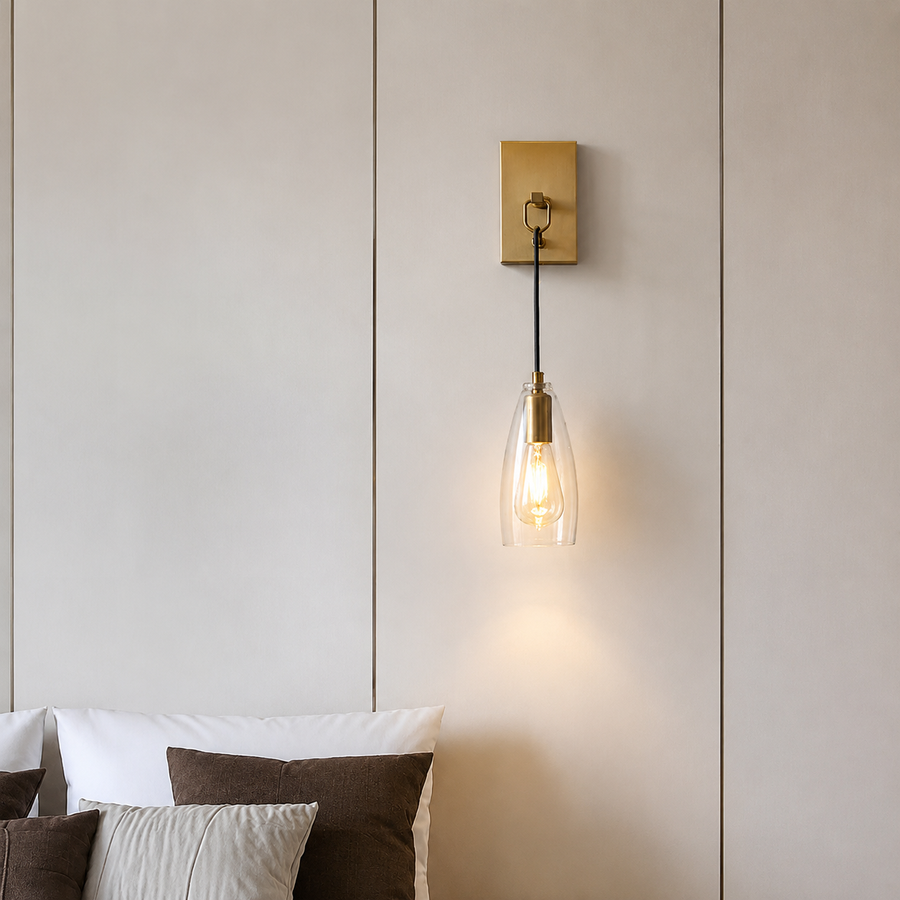 A modern wall-mounted sconce with a square gold brass backplate, black cord, and a clear glass cone shade housing a warm glowing Edison bulb, shown lit on a beige paneled wall above a bed.