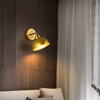 Gold metal wall light with dome shade casting warm glow styled beside bed creating cozy ambient lighting for bedroom decor.