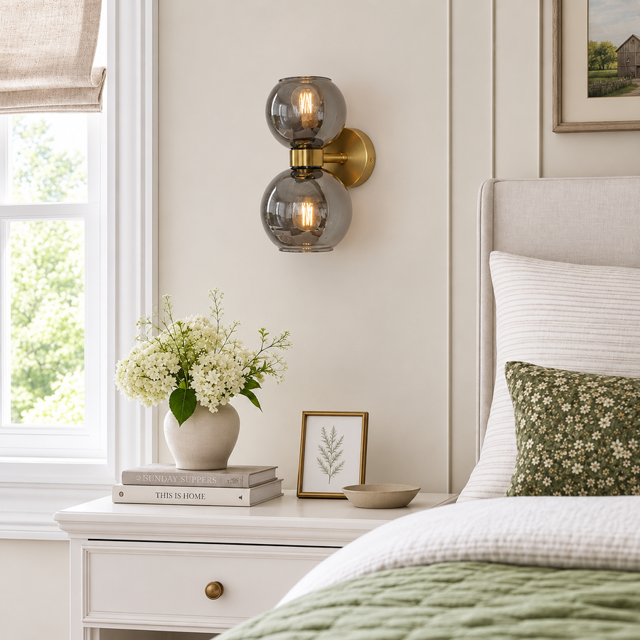 A modern wall-mounted sconce with two stacked smoky glass globes and a brushed brass backplate, lit with warm bulbs, mounted on a cream bedroom wall above a white nightstand.