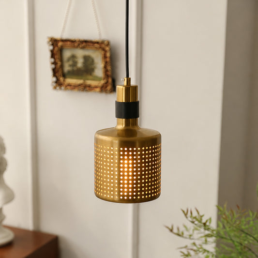 A modern pendant light with a brushed brass cylindrical shade featuring a perforated pattern, suspended by a black cord against a white paneled wall.
