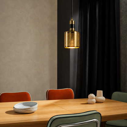 A modern cylindrical pendant light with a textured gold mesh shade hangs over a wooden dining table in a styled interior setting with chairs and neutral decor.