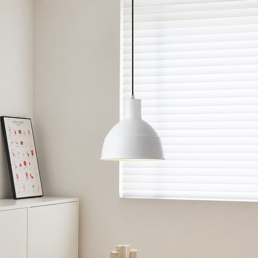 A modern white pendant light with a matte finish hangs from a black cord in front of white window blinds in a bright, minimalist room.