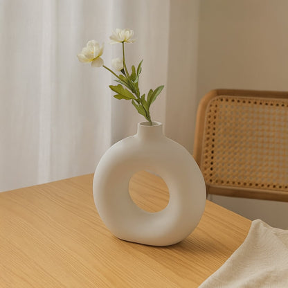 Main lifestyle image showing a white ceramic decorative vase with circular cutout placed on a wooden dining table styled with flowers in a modern home setting.