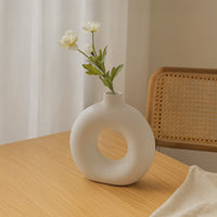 Main lifestyle image showing a white ceramic decorative vase with circular cutout placed on a wooden dining table styled with flowers in a modern home setting.