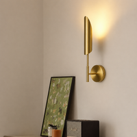 A modern wall-mounted sconce with a brushed gold finish, shown lit on a beige wall above a console table with a framed print and a speaker.