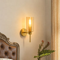 Modern gold brass wall sconce with amber glass cylinder shade glowing in a cozy bedroom nightstand setup corner.