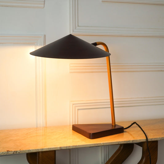 A modern table lamp with a matte black conical shade, a warm orange-brown curved metal stem, and a dark wood base, illuminated on a light marble tabletop against a cream paneled wall.