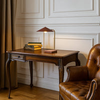 Modern brass desk lamp with angular shade illuminating a wooden desk beside leather chair in elegant study room.