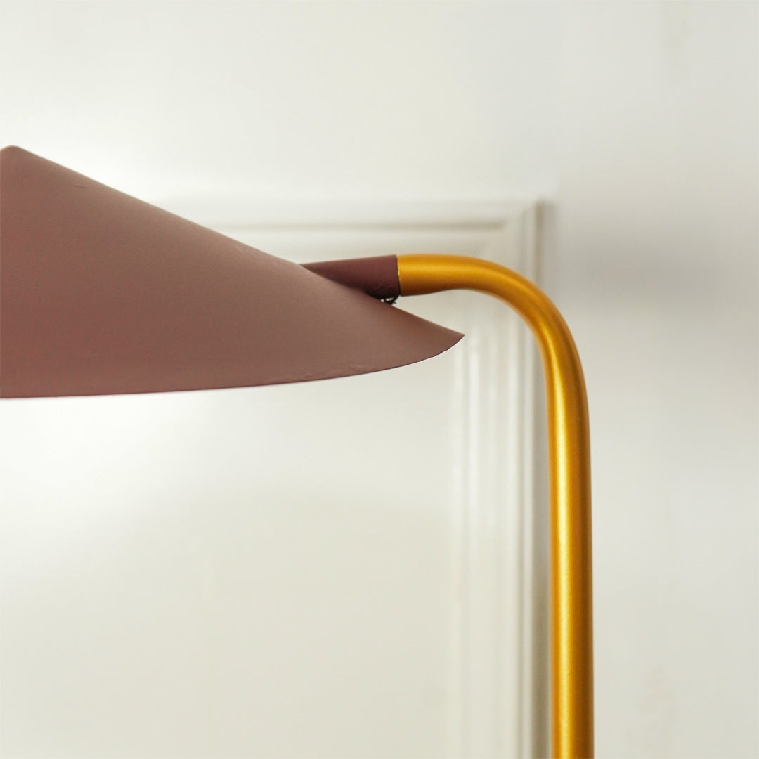 Close-up of a modern floor lamp with a curved gold metal stem and a wide, tapered dusty rose-brown shade, shown against a white wall and door frame.
