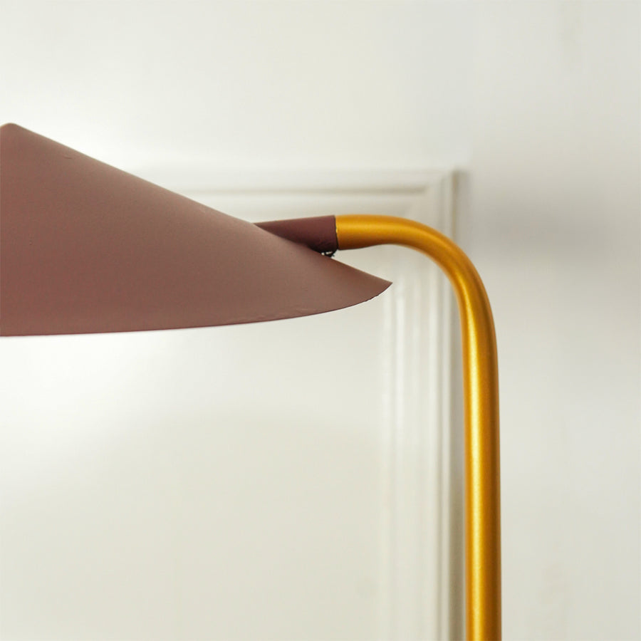 Close-up of a modern floor lamp with a curved gold metal stem and a wide, tapered dusty rose-brown shade, shown against a white wall and door frame.