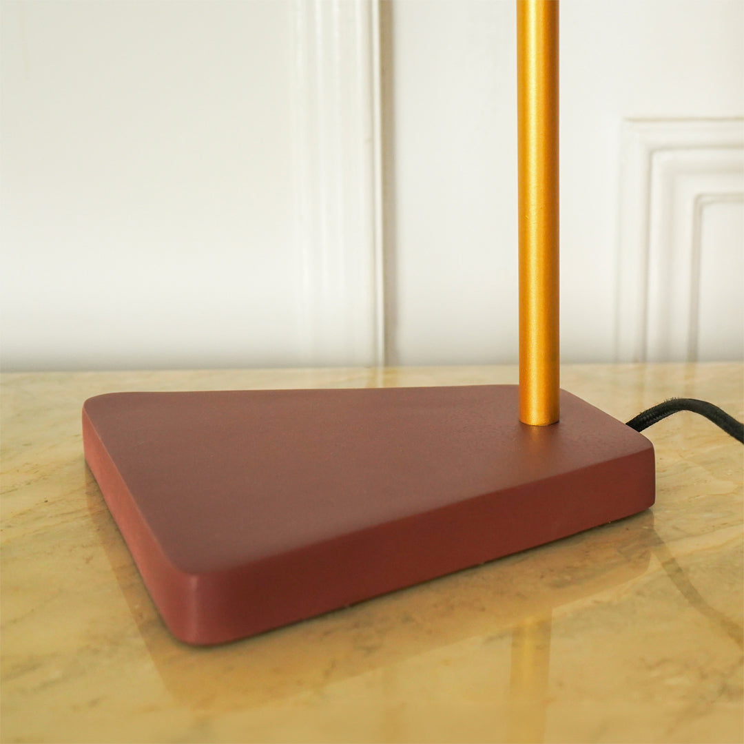 A modern table lamp with a matte terracotta-colored rectangular base and a slim metallic gold pole, shown on a polished marble tabletop against a white paneled wall.