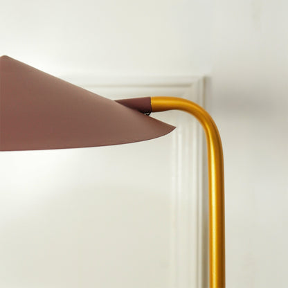 Close-up of a modern floor lamp with a curved gold metal stem and a wide, tapered dusty rose-brown shade, shown against a white wall and door frame.