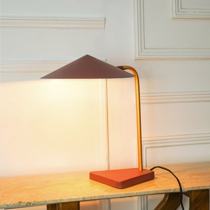 A modern table lamp with a terracotta orange rectangular base, a gold-finished curved metal stem, and a dark brown conical shade, shown illuminated on a light wood table against a white paneled wall.