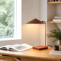 A modern table lamp with a dark reddish-brown conical shade, a gold-toned curved stem, and a square orange-brown base sits on a wooden desk in a bright room. An open book, a small plant in a glass vase, and stacked books are nearby.