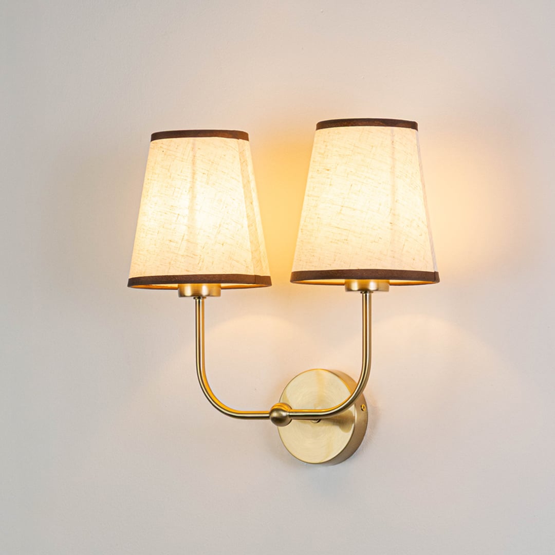 Gold metal double wall sconce with cream textured shades illuminated on a bright white background for clear visibility.