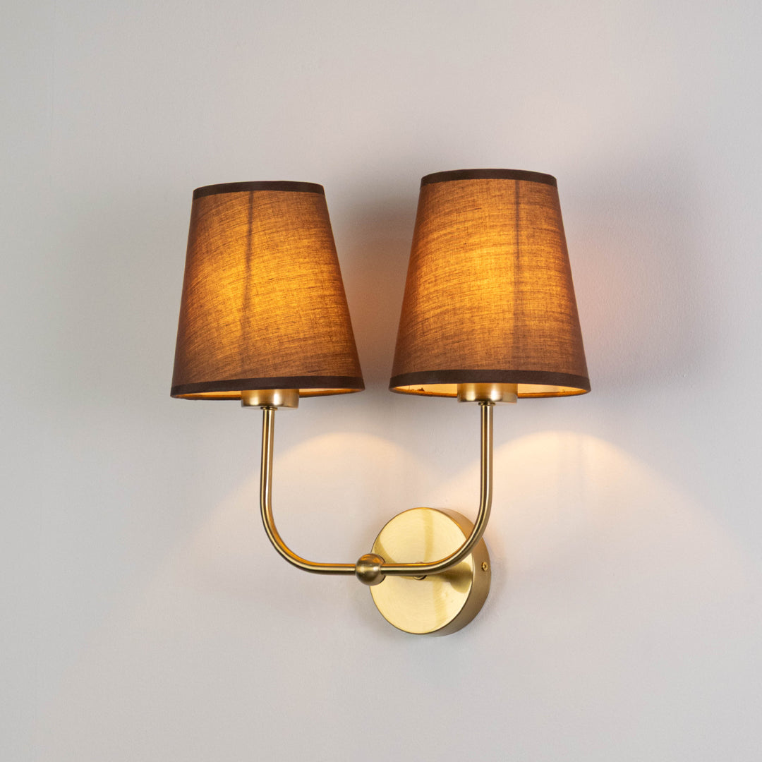 Contemporary wall light in gold metal finish with dual fabric shades for bedroom or living room ambient lighting decor white background.