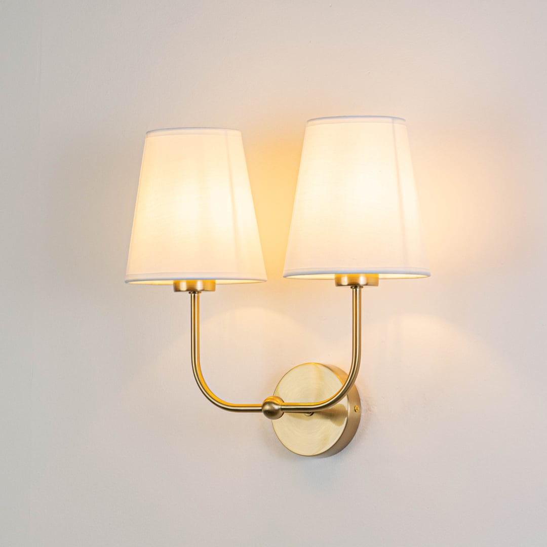 Gold metal double wall sconce with white fabric shades illuminated on a bright white background for clarity.