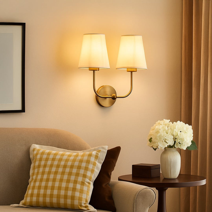 Gold metal double wall sconce with white fabric shades adding soft warm lighting to a modern home interior.