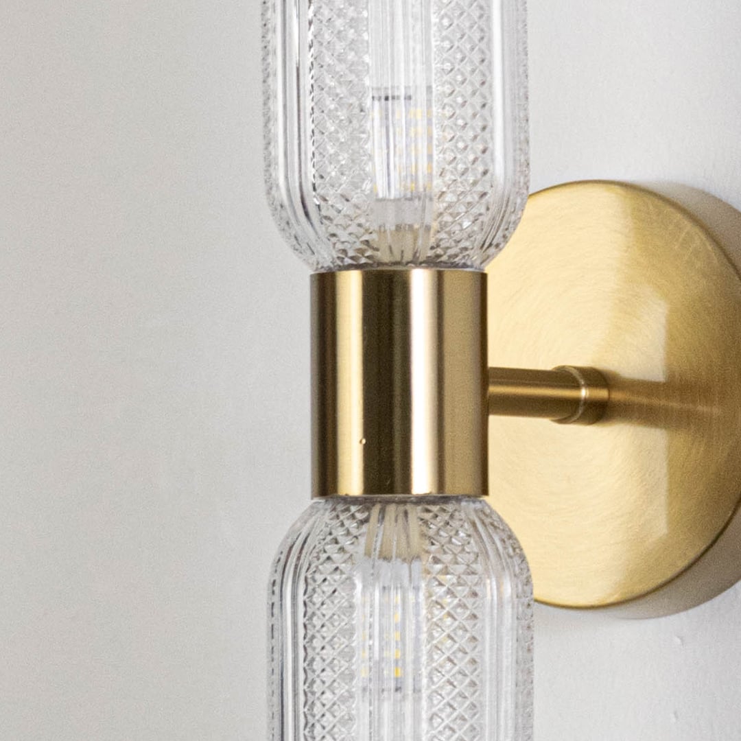 Closeup of modern gold wall sconce with dual textured glass tube shades and round metal base showing elegant craftsmanship details.