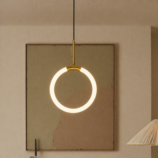 A modern circular LED pendant light with a gold-finished top cap and black cord, hanging in front of a textured beige wall and a framed artwork.