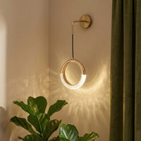 Modern gold brass curved arm hanging ring LED wall sconce casting a radiant geometric glow on a corner wall next to a plant.