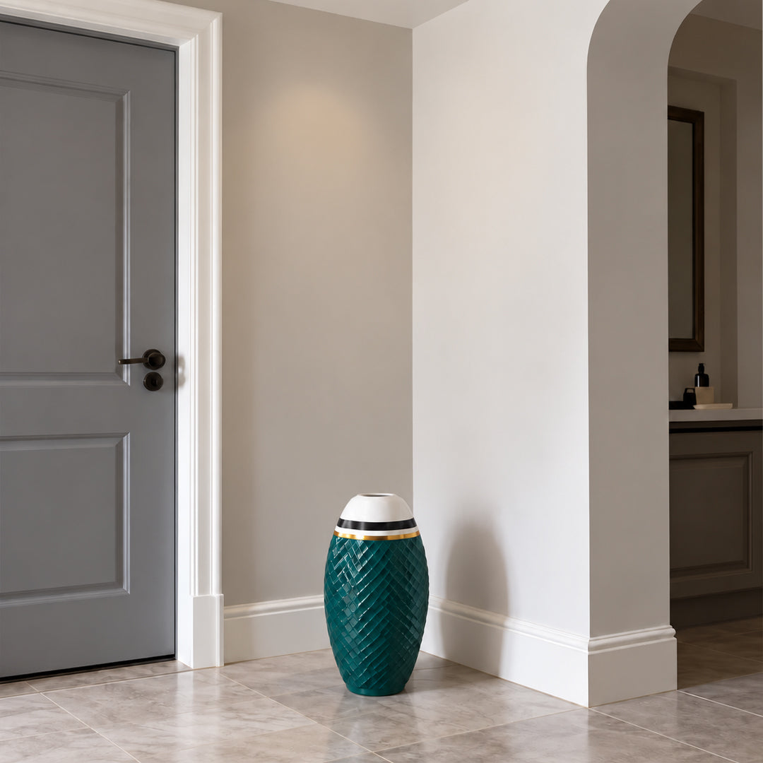 A decorative teal ceramic drum vase with a textured chevron pattern, a white top, and gold band accents, placed on a light stone tiled floor in a modern hallway.
