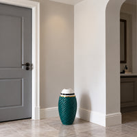 A decorative teal ceramic drum vase with a textured chevron pattern, a white top, and gold band accents, placed on a light stone tiled floor in a modern hallway.