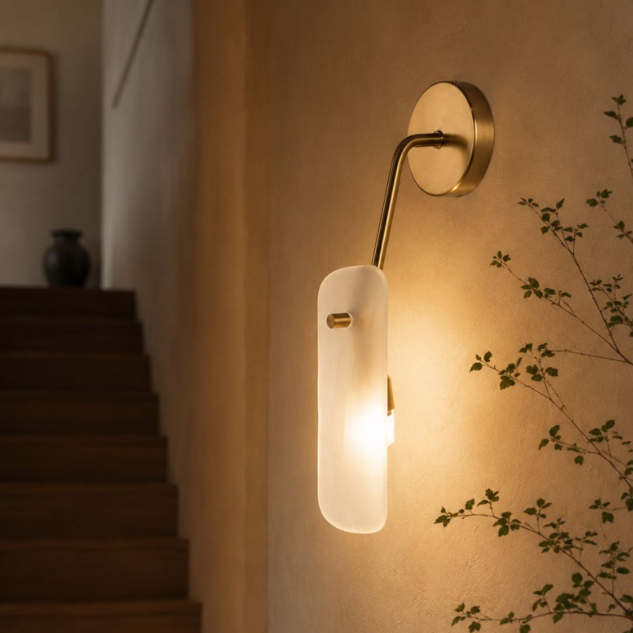 A modern wall sconce light fixture with a vertical frosted glass shade and a curved brass arm, mounted on a beige wall next to a staircase.