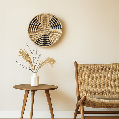 Handwoven round wall basket Decor displayed on a soft neutral wall above a wooden side table with dried grass and a woven chair creating a calm earthy and minimalist interior setting.