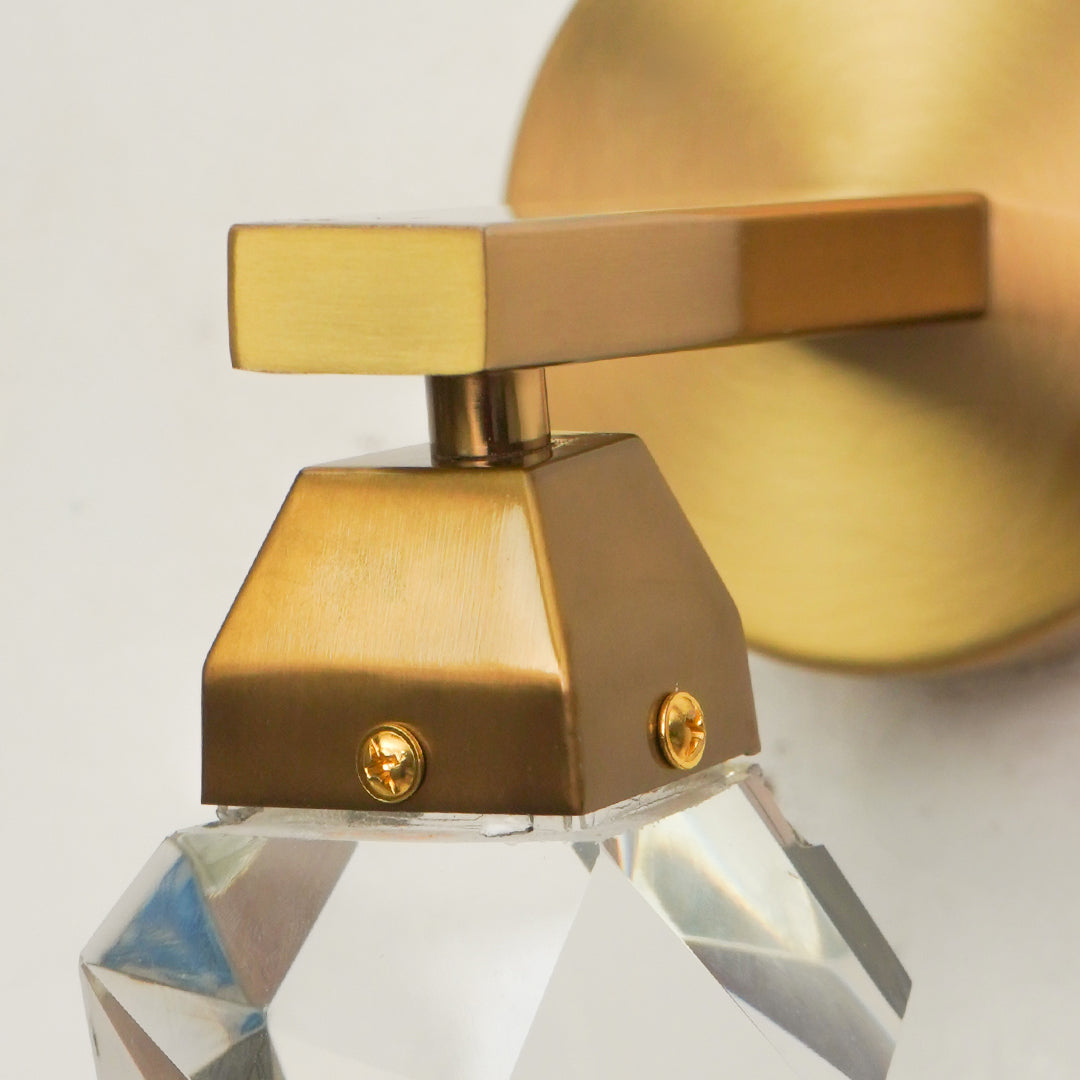 A close-up of a modern soap dispenser with a brushed brass-finish metal body and a clear faceted crystal base, set against a light neutral background.