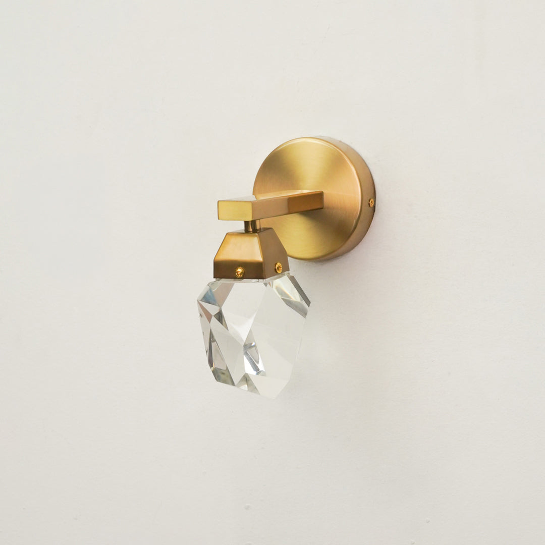 A modern wall-mounted light fixture with a brushed gold circular backplate, a geometric gold metal arm, and a faceted clear crystal shade, shown against a plain off-white wall.