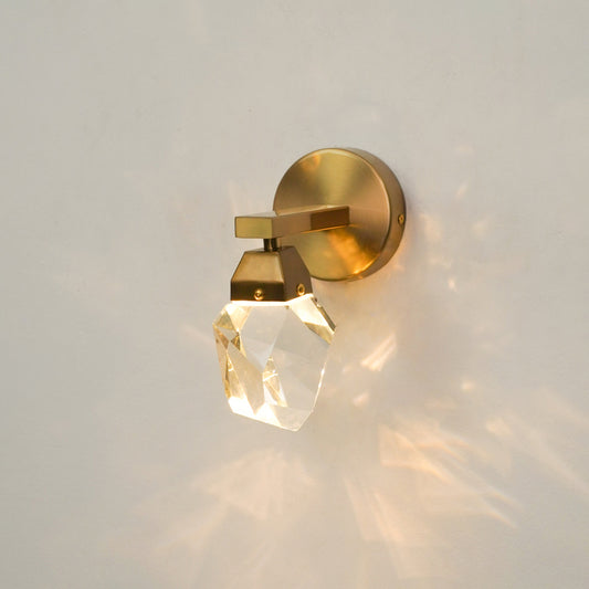 A modern wall-mounted light fixture with a brushed gold circular backplate, a geometric gold metal arm, and a faceted clear crystal shade, shown lit against a plain light wall with warm light reflections.