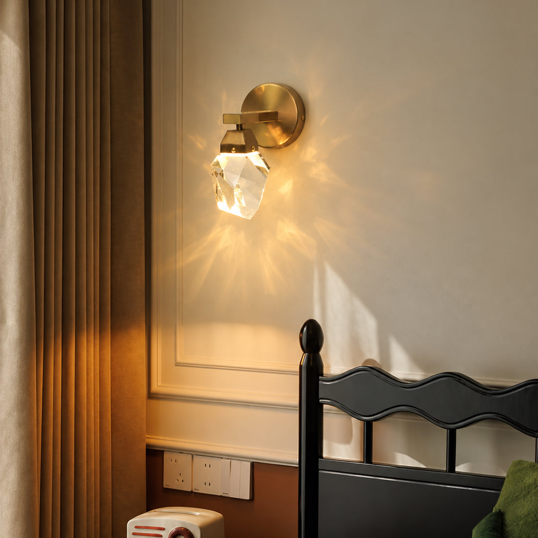 A brass wall-mounted sconce with a faceted clear crystal shade, glowing warm light on a beige wall above a dark wooden headboard.