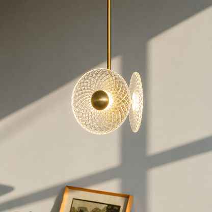 A modern pendant light with a textured, woven spherical glass shade and a brushed brass rod, casting a soft shadow on a light gray wall.