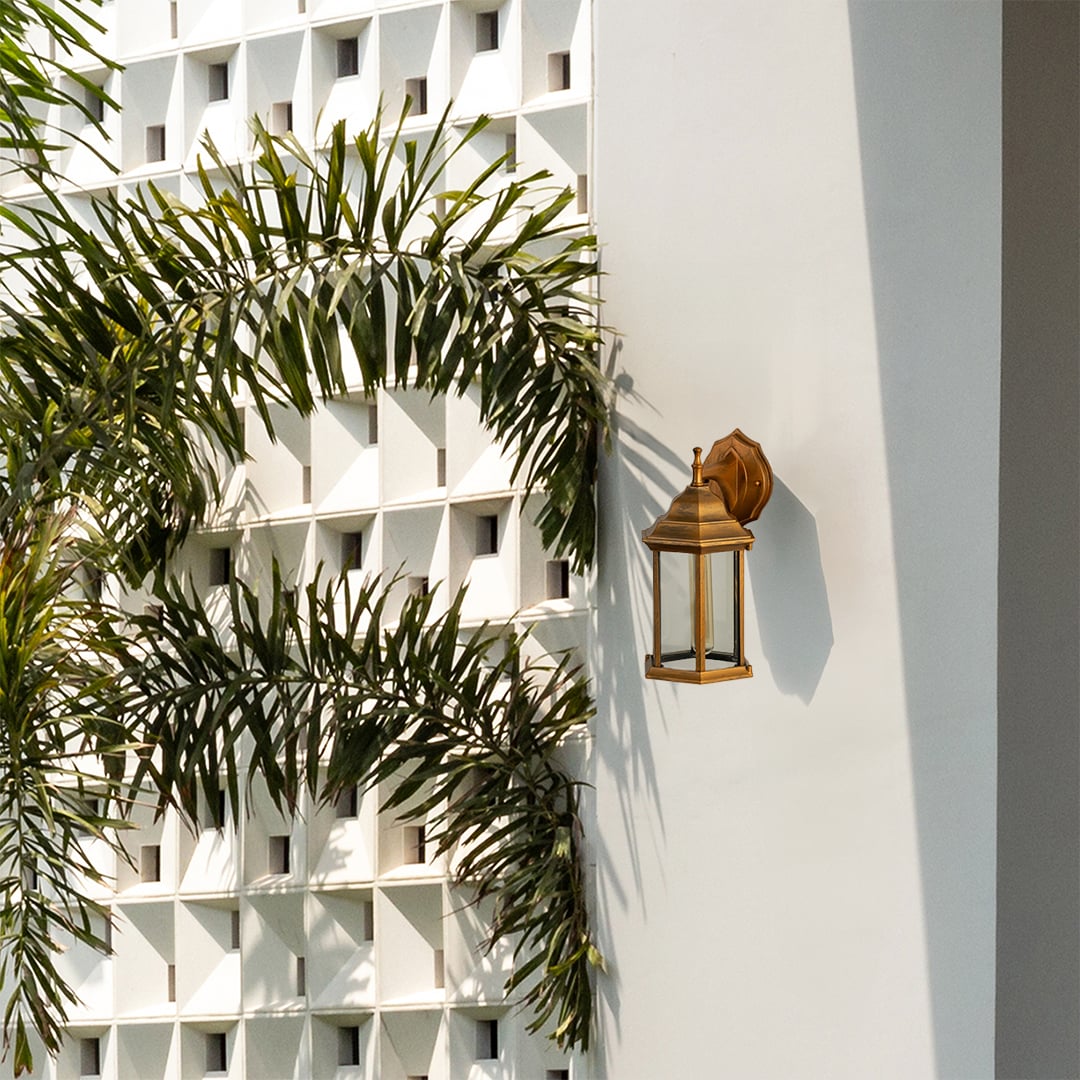 Antique brass outdoor wall lantern lifestyle image with clear glass panels mounted on white villa wall with palm trees.