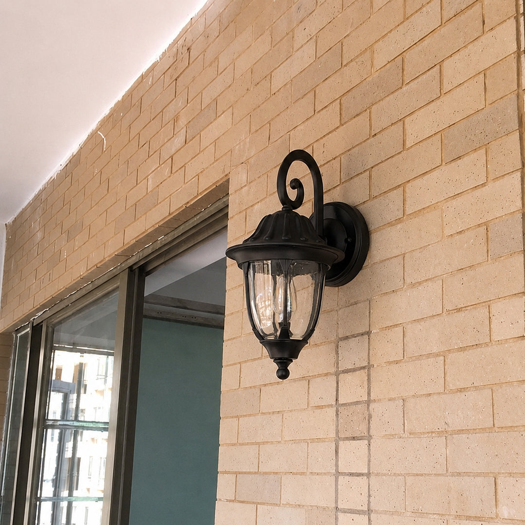 Black metal outdoor wall light lifestyle image with clear glass shade mounted on a tan brick entryway patio wall.